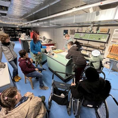 Sechs Schüler: innen und eine Lehrerin hören dem Museumsmitarbeiter zu. Sie stehen um einen großen Computer in der Mitte des Raumes herum