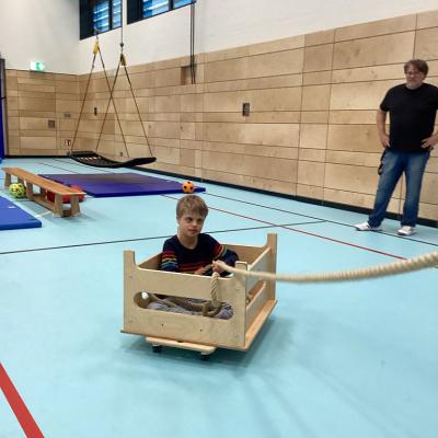 Ein Junge sitzt in einer Holzkiste auf Rollen. Der Junge zieht sich mit Hilfe eines dicken Seils selbstständig in die Richtung des Fotografens