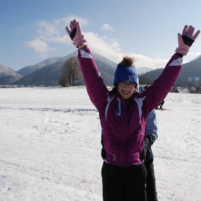 Marija steht, während sie von Frau Fecke gehalten wird. Sie reckt die Arme in die Höhe und lächelt in die Kamera. Im Hintergrund sieht man ein großes Schneefeld und die schöne Landschaft mit dem guten Wetter.