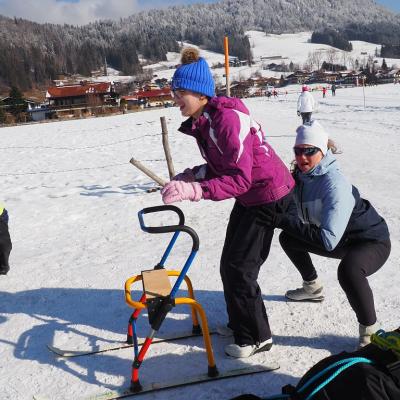 Marija steht vor ihrem Skilator und macht beim Aufwärmen mit. Frau Fecke ist hinter ihr in der Hocke. Im Hintergrund sieht man ein großes Schneefeld und die schöne Landschaft mit dem guten Wetter.