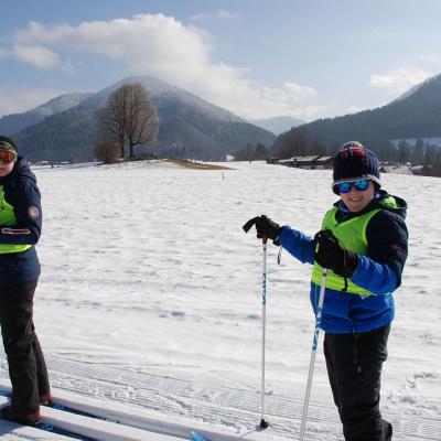 In der Loipe stehen Christopher und Josua. Sie drehen sich zur Kamera und lächeln. Im Hintergrund sieht man ein großes Schneefeld und die schöne Landschaft mit dem guten Wetter.