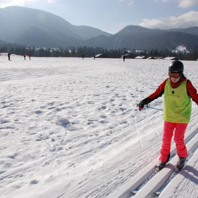 Lara läuft in der Loipe. Ihre Arme balancieren im Schwung das Gleichgewicht. Im Hintergrund sieht man ein großes Schneefeld und die schöne Landschaft mit dem guten Wetter.