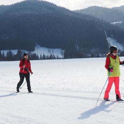 Lara und Frau Gauch laufen von links nach rechts durchs Bild auf der Loipe. Im Hintergrund sieht man ein großes Schneefeld und die schöne Landschaft mit dem guten Wetter.