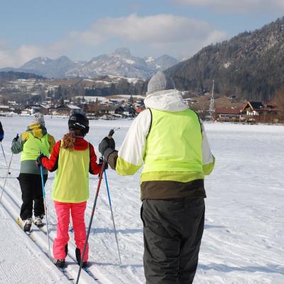 Man sieht eine Gruppe von Schüler:innen in der Loipe von hinten laufen. Es sind Christopher, Josua, ein Schüler aus Stuttgart, Lara und Julian. Im Hintergrund sieht man ein großes Schneefeld und die schöne Landschaft mit dem guten Wetter.
