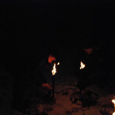 Im Dunklen erkennt man gerade so Christophers Gesicht. Er beugt sich nach unten und steckt eine Fackel in den Schnee. Drei Flammen brennen in der Schwärze.