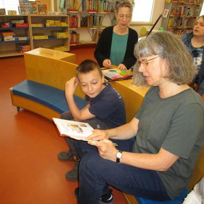 Ein Junge sitzt links von seiner Lehrerin, die ein Buch in der Hand hält. Er liest mit ihr. Dahinter sind zwei weitere Frauen in der Bücherei zu sehen.