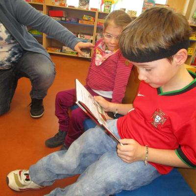 Ein Junge im roten Trikot liest ein Kinderbuch. Ein Mädchen sitzt daneben und schaut interessiert zu.