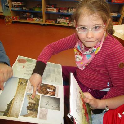 Ein Mädchen sitzt auf der Bank und zeigt auf ein Bild in einem Sachbuch über Pferde. Im Hintergrund Spieleregale.