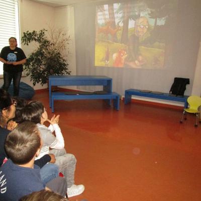 Kinder und Erwachsene schauen an die Wand, auf die ein Bilderbuchkino projiziert wird. Die Büchereimitarbeiterin liest im Stehen die Geschichte vor.