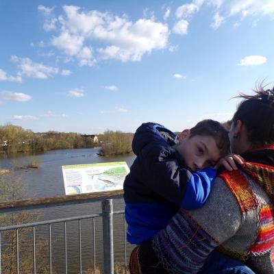 Eine Frau und ein Junge sind auf einer Aussichtsplattform. Die Frau trägt den Jungen und steht mit dem Rücken zur Kamera. Der Junge schaut in Richtung Kamera. Im Hintergrund sind man weitläufig einen Fluss und viele Bäume und Büsche.  