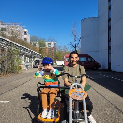 Ein Junge und ein Mann sitzen nebeneinander auf einem orangenen Fahrrad.  Die Sonne scheint. Beide lachen, der Junge hebt die Hände
