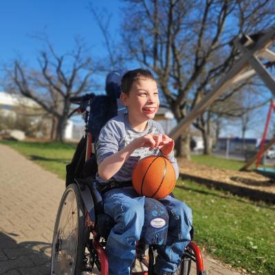 Ein Junge im Rollstuhl schaut breit lachend nach vorne. Auf seinem Schoß liegt ein Basketball. Die Sonne scheint. 