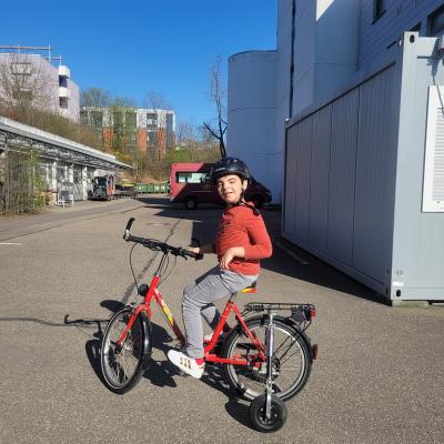 Ein Junge sitzt auf einem Fahrrad mit Stützrädern. Er lächelt in die Kamera. Die Sonne scheint. 