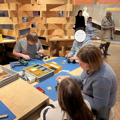 Drei Kinder mit einer Frau sie sind im Bauzimmer der Experimental. Sie sitzen am Tisch und bauen Autos. Sie benutzen Schrauben, Schraubendreher, Hammer und eine Schere.