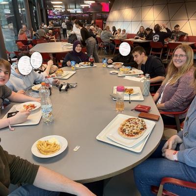 An einem großen Tisch sitzen 2 Frauen, 1 Mann und 6 Jugendliche. Sie essen Pommes mit Burger, Nuggets, Pizza und Gulasch mit Nudeln. Sie sitzen in der Kantine der Experimenta Alle freuen sich und lächeln in die Kamera.  