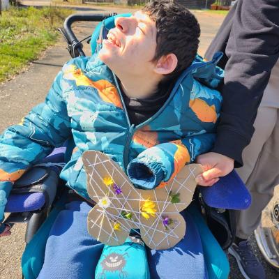 Schüler im Rollstuhl ,hat seinen Schmetterling auf dem Schoss , dabei lächelt er in die Sonne , der Rollstuhlfahrer ist in blau gekleidet , der Rollstuhl ist Türkis, die Sonne Strahlt 