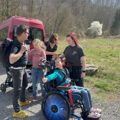 Drei Betreuer, zwei Rollstuhlfahrer eine Läuferin sind aus dem Roten Bus ausgestiegen und laufen Richtung Naturpfad