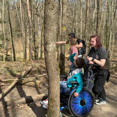 Ein Rollstuhlfahrer schaut in den Wald uns sucht das Tier, die Betreuerin steht hinter ihm und sucht auch , im Hintergrund zeigt ein Betreuer einer Schülerin wo das Tier sich versteckt hat 