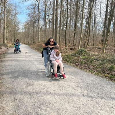 Eine Rollstuhlfahrern fährt auf dem Waldweg die Betreuerin schiebt sie den Berg hoch 
