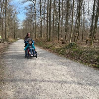 Ein Rollstuhlfahrer fährt auf dem Waldweg die Betreuerin schiebt ihn  den Berg hoch