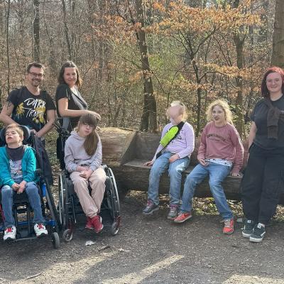 Links ein Rollstuhlfahrer , dahinter ein Betreuer, daneben eine Rollstuhlfahrerin dahinter eine Betreuerin auf der Baum Bank eine sitzende Schülerin , daneben eine Betreuerin alle lachen in die Kamera 