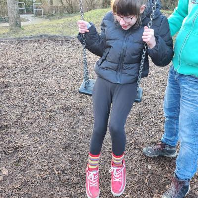 Eine Schülerin sitzt auf der Schaukel und lacht, sie hat eine Schwarze dicke Winterjacke an, eine schwarze Leggings ,bunte Ringel Socken, rot-weiße schuhe
