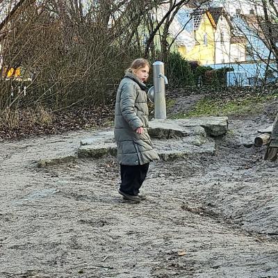 Schülerin steht im Sand mit einer  Olivfarbenden Wintermantel ,schwarze Hose , schau erstaunt in die Kamera