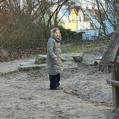 Schülerin steht im Sand mit einer  Olivfarbenden Wintermantel ,schwarze Hose , schau erstaunt in die Kamera