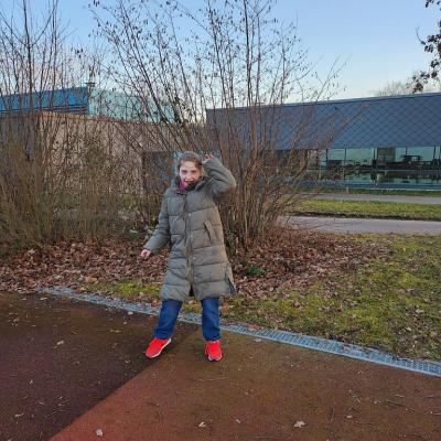 Schülerin auf dem roten Sportplatz, der Boden ist rot, im Hintergrund sind Sträucher und die Schwimmhalle. 