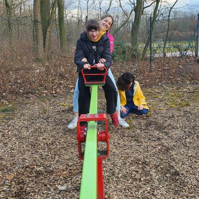 Schülerin sitzt lachend auf der Wippe dahinter eine Betreuerin , im Hintergrund Seite sitzt eine Schülerin auf dem Boden und wartet bis sie wieder Wippen darf