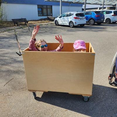 Zwei Schülerinnen sitzen im Holzwagen, man sieht nur zwei Köpfe , eine jubelt mit nach oben gestreckten Armen und die andere schaut über die kante