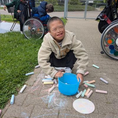 Ein Junge hockt auf dem Boden. Vor ihm ist ein Leerer Eimer, daneben überall Kreidestifte verteilt. Der Junge lächelt. Um ihn herum auf dem Boden wurde gemalt. 