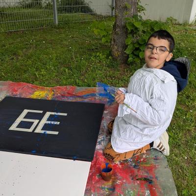 Ein Junge kniet vor einer Leindwand. Mit einem Pinsel streicht er blaue Farbe durch ein Sieb.  