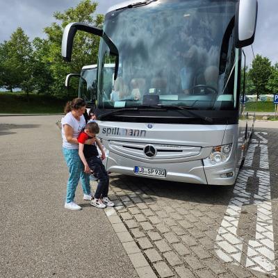 Man sieht einen großen grauen Reisebus von vorne. Vor dem Reisebus laufen eine Frau und ein Junge. Die Frau steht hinter dem Jungen und hilft ihm 