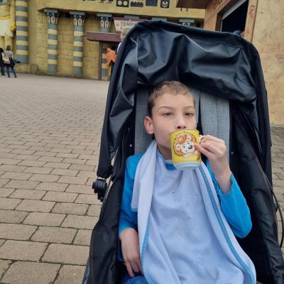 Ein Junge sitzt in einem Buggy und trinkt aus einer Tasse mit einem Löwen darauf.  Im Hintergrund ist ägyptisch angehauchtes Gebäude 