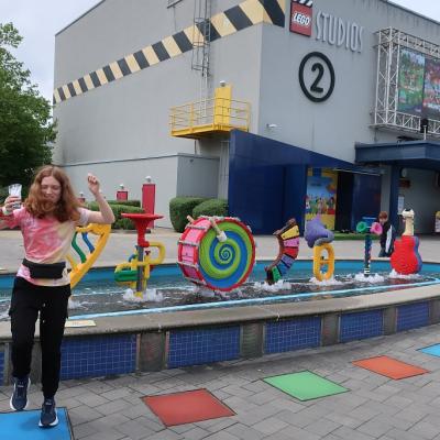Ein Mädchen steht vor einem Springbrunnen und Springt in die Luft. Im Brunnen sind Figuren aus Lego 