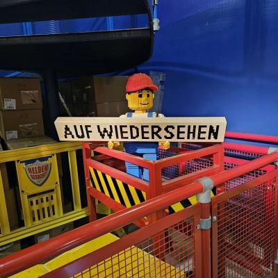 Ein Männchen aus Lego hält ein Schild in die Luft auf dem „Auf Wiedersehen“ steht. 