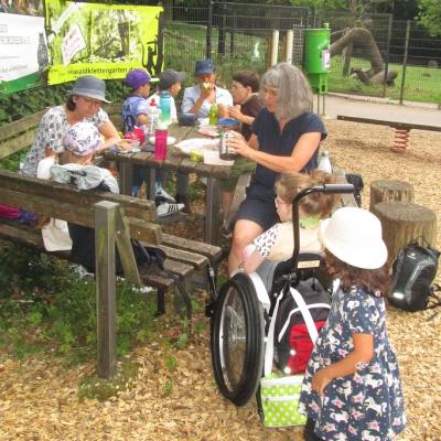 3 Erwachsene und 6 Kinder mit Behinderung im Grundschulalter vespern an einem Tisch mit Bänken im Wildpark Pforzheim. Alle sind sommerlich gekleidet.
