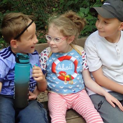 2 Jungen und ein Mädchen, alle im Grundschulalter, auf einer Bank, das strahlende Mädchen in der Mitte