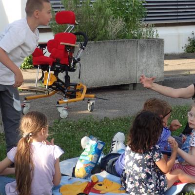 4 Kinder und eine Frau sitzend auf einer Decke im Gras vor dem Schulgebäude, ein Junge stehend. Chipstüte in der Mitte