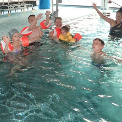 4 Kinder und 2 Frauen im Schulschwimmbad, teilweise mit Schwimmflügeln. Die meisten strecken die Hände nach oben und lachen.