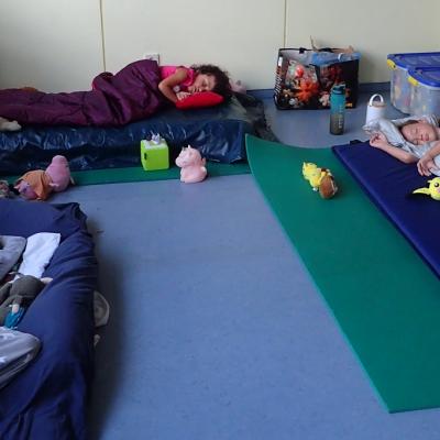 3 Kinder liegen auf Matten in Schlafsäcken oder Decken u-förmig angeordnet in der Spieleecke des Klassenzimmers und schlafen