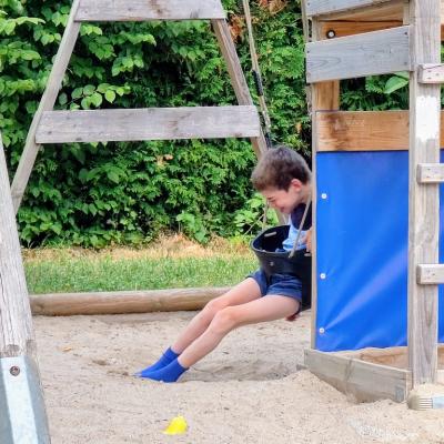 Schüler sitzt in einer Schaukel , lacht dabei weil er alleine Schaukeln kann , Füße sind im Sand zum abstoßen