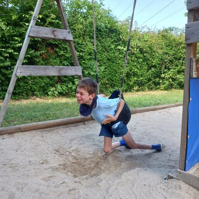 Schüler ist in der Schaukel nach vorne gebeugt , strahlt, lacht , die Füße sind im Sand , im Hintergrund sind die Sträucher im prachtvollem grün, Himmel ist blau 