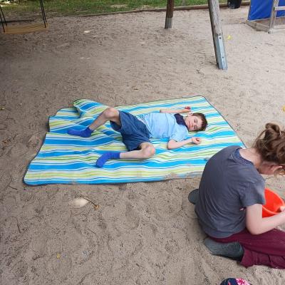 Schüler liegt auf einer grün, blau, weiß gestreiften decke im Sand , Schülerin sitzt daneben und buddelt im Sand 