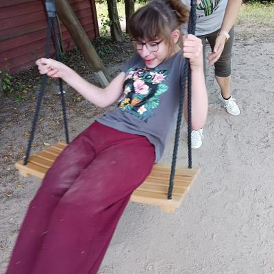 Schülerin sitzt auf einer Holzschaukel, wird von einer Betreuerin geschaukelt, Schülerin hat eine rote Hose an , ein graues Shirt, eine rote Brille