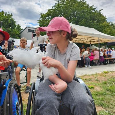 Schülerin im Rollstuhl bekommt eine Zuckerwatte hingehalten, sie Freut sich, weiß aber nicht was das ist. Im Hintergrund sind viele andere Gäste vom Fest 