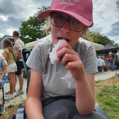 Schülerin mit rosa farbende Cape , roter Brille, graues Shirt , graue Hose , hält in der Hand die weise Zuckerwatte und probiert vorsichtig mit dem Mund 