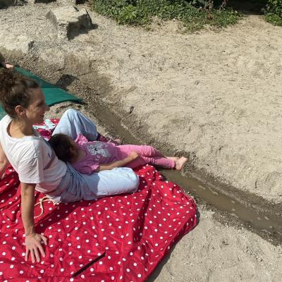 Mädchen sitzt mit Lehrerin auf der Picknickdecke am Bachlauf und streckt die nackten Füße ins Wasser