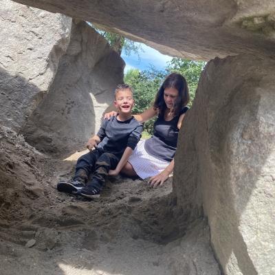 Junge sitzt mit seiner Lehrerin in einer Höhle aus Steinen (im Schatten) auf dem Wasserspielplatz. Er lacht fröhlich und freut sich darüber, dass er mit dem Sand spielt.
